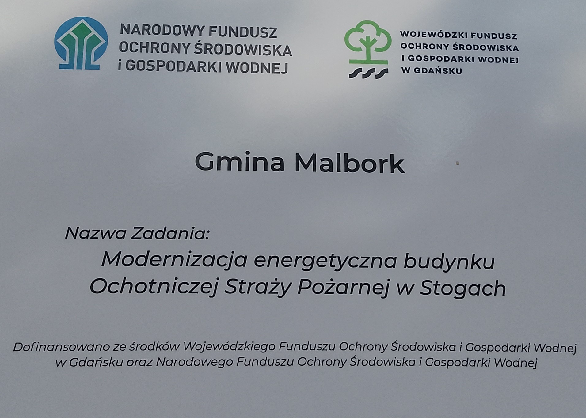  Tablica informacyjna dotycząca zadania &bdquo;Modernizacja energetyczna budynku Ochotniczej Straży Pożarnej w&nbsp;Stogach&rdquo;. Zawiera logotypy i&nbsp;informację o&nbsp;dofinansowaniu ze środk&oacute;w Wojew&oacute;dzkiego Funduszu Ochrony Środowiska i&nbsp;Gospodarki Wodnej w&nbsp;Gdańsku oraz&nbsp;Narodowego Funduszu Ochrony Środowiska i&nbsp;Gospodarki Wodnej.  Na zdjęciu widoczna jest jasna tablica informacyjna. W jej g&oacute;rnej części znajdują się dwa logotypy  Po lewej stronie znajduje się logo Narodowego Funduszu Ochrony Środowiska i&nbsp;Gospodarki Wodnej: niebieski okrąg z&nbsp;trzema zielonymi, geometrycznymi kształtami przypominającymi drzewa, obrysowanymi jasną linią. Obok widnieje pełna nazwa Funduszu zapisana czarnymi literami.  Po prawej stronie znajduje się logo Wojew&oacute;dzkiego Funduszu Ochrony Środowiska i&nbsp;Gospodarki Wodnej w&nbsp;Gdańsku: zielone, stylizowane drzewo z&nbsp;zaokrągloną koroną i&nbsp;pniem, pod kt&oacute;rym umieszczono trzy czarne, faliste elementy symbolizujące wodę. Obok widnieje pełna nazwa Funduszu zapisana czarnymi literami.  Pośrodku umieszczono napis &bdquo;Gmina Malbork&rdquo;, a&nbsp;poniżej nazwę zadania: &bdquo;Modernizacja energetyczna budynku Ochotniczej Straży Pożarnej w&nbsp;Stogach&rdquo;. W dolnej części tablicy znajduje się informacja o&nbsp;dofinansowaniu zadania ze środk&oacute;w obu funduszy. 